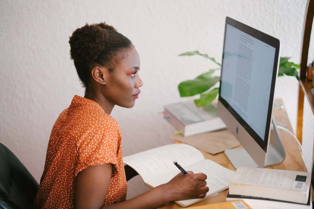 adult woman looking at computer screen while taking notes on notepad